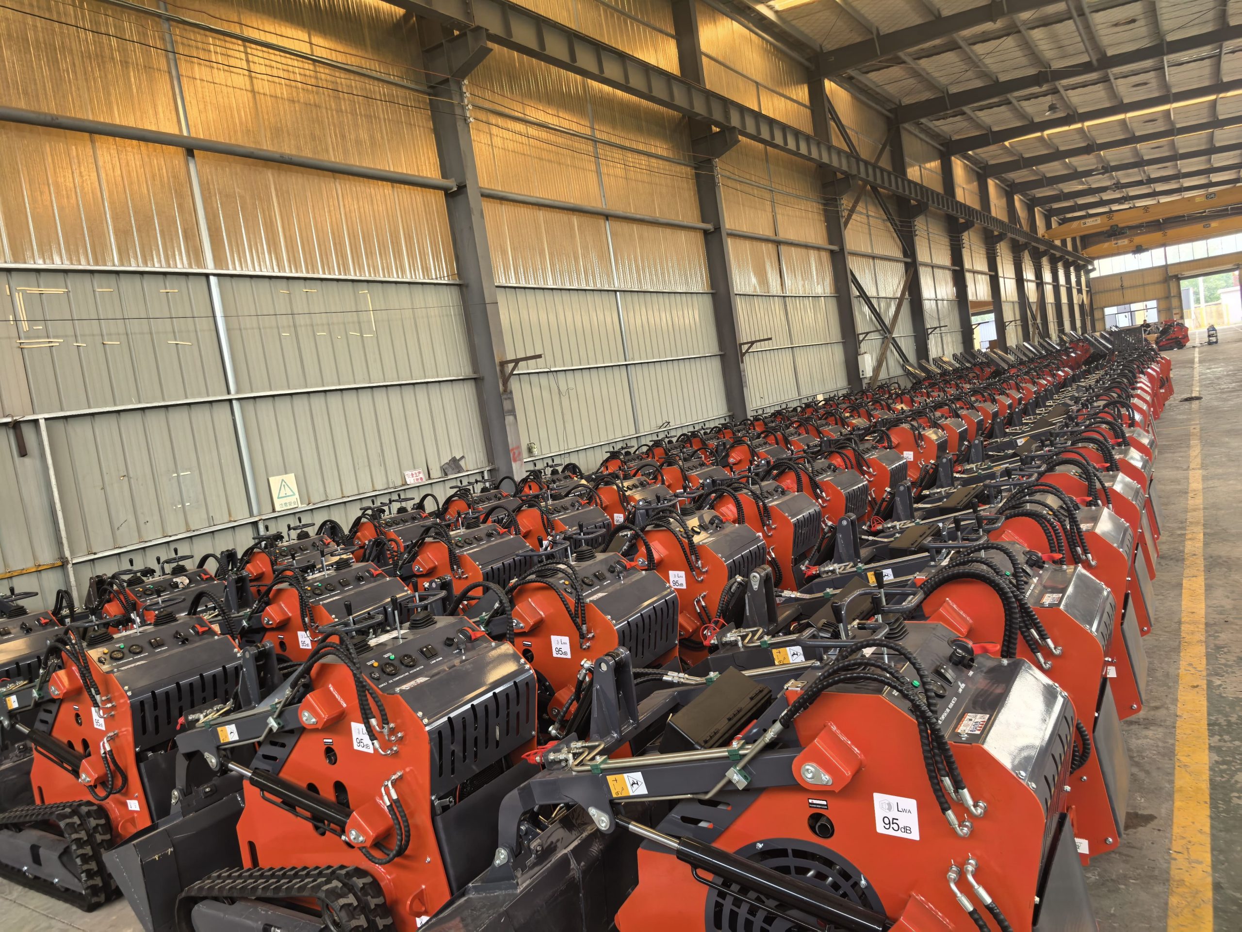 Rows of orange tracked mini skid steer loaders lined up in a large steel-framed factory warehouse